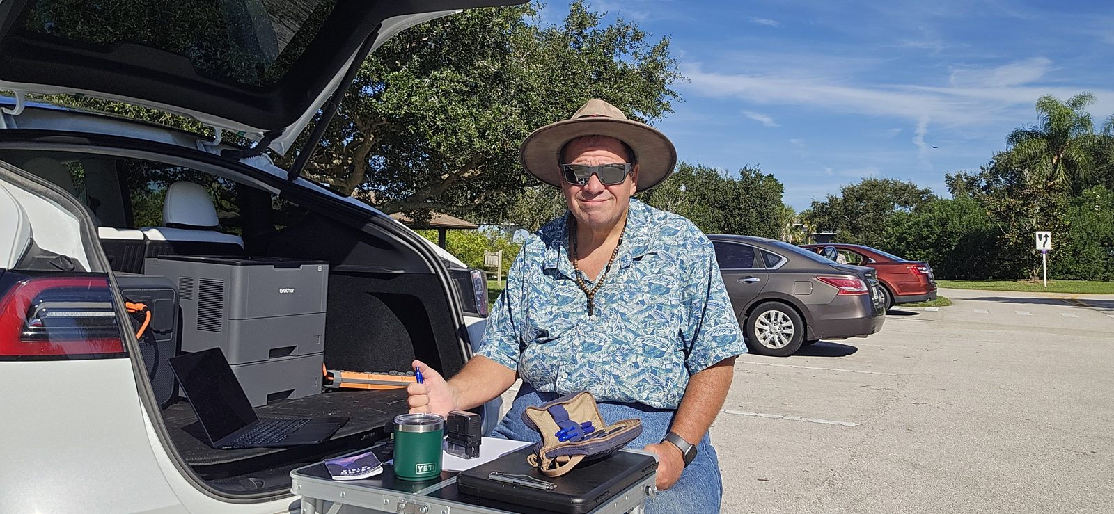 Man in a hat seated at a folding table behind an open white Tesla hatch outdoors